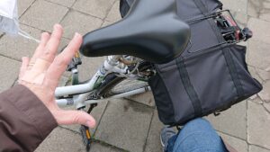 Ein Fahrrad von oben, mit zwei Packtaschen am Gepäckträger, das von einer Hand und einem Knie am Umfallen gehindert wird.