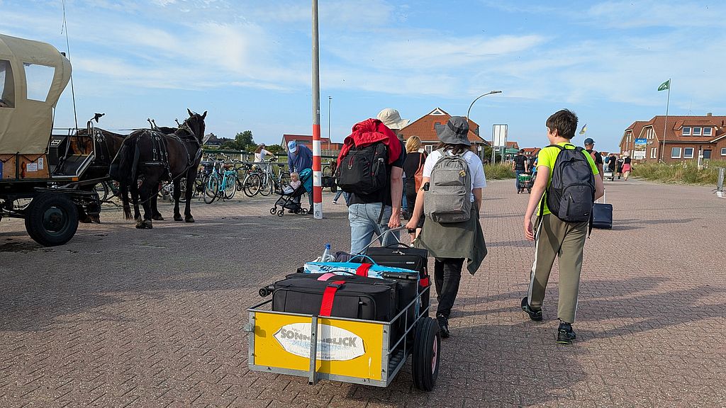 Drei Personen ziehen einen mit Koffern beladenen Handwagen, im Hintergrund sieht man Fahrräder und eine Pferdekutsche