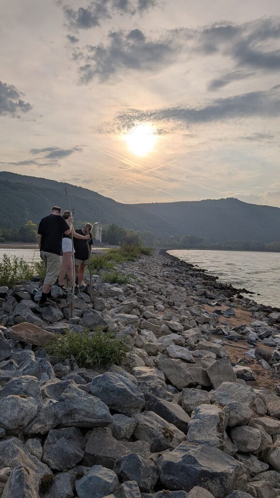 Drei Leute stehen auf einem steinigen Wall mitten im Rhein, im Hintergrund der Mäuseturm, die bewaldeten Hänge des Hunsrück und eine niedrigstehende Sonne in einem leicht bewöltken Himmel.